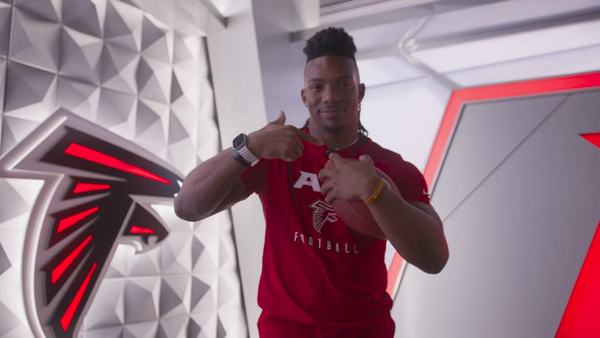 Bijan Robinson in Atlanta Falcons team apparel holding a football beside the Falcons logo wall