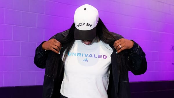 A person wearing a white Unrivaled T-shirt with the adidas logo and a Bay Area cap against a purple wall